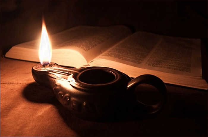 A lit oil lamp sits beside an open book in a dimly lit setting, casting a warm glow.