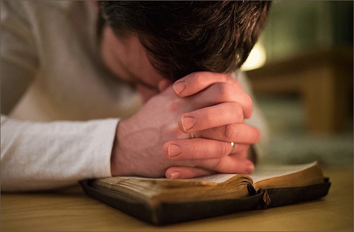 A person with clasped hands leaning over an open book, appearing to pray or reflect.