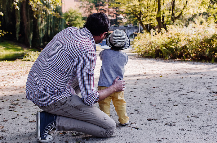 A father kneels next to his young child, who is looking away, in a park setting with trees and a pathway.