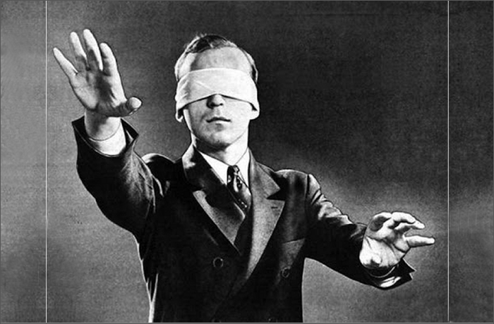 A black and white image of a man in formal attire with a blindfold, reaching forward with outstretched hands.