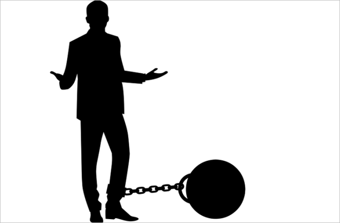 Silhouette of a man in a suit looking confused with his arms raised, attached to a large ball and chain at his feet.