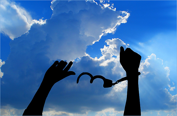 Silhouetted hands breaking free from handcuffs against a bright blue sky with clouds.