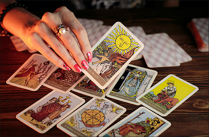 A person's hand with stylish nails holding a tarot card featuring a sun symbol, surrounded by various other tarot cards spread out on a wooden surface.