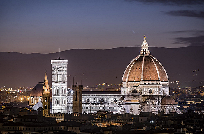 A panoramic view of the Florence Cathedral at night, highlighting its iconic dome and bell tower against a twilight sky.
